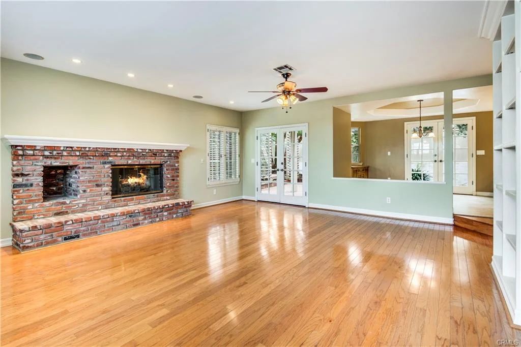 Large living room with wooden floor, brick fireplace, ceiling fan, and windows