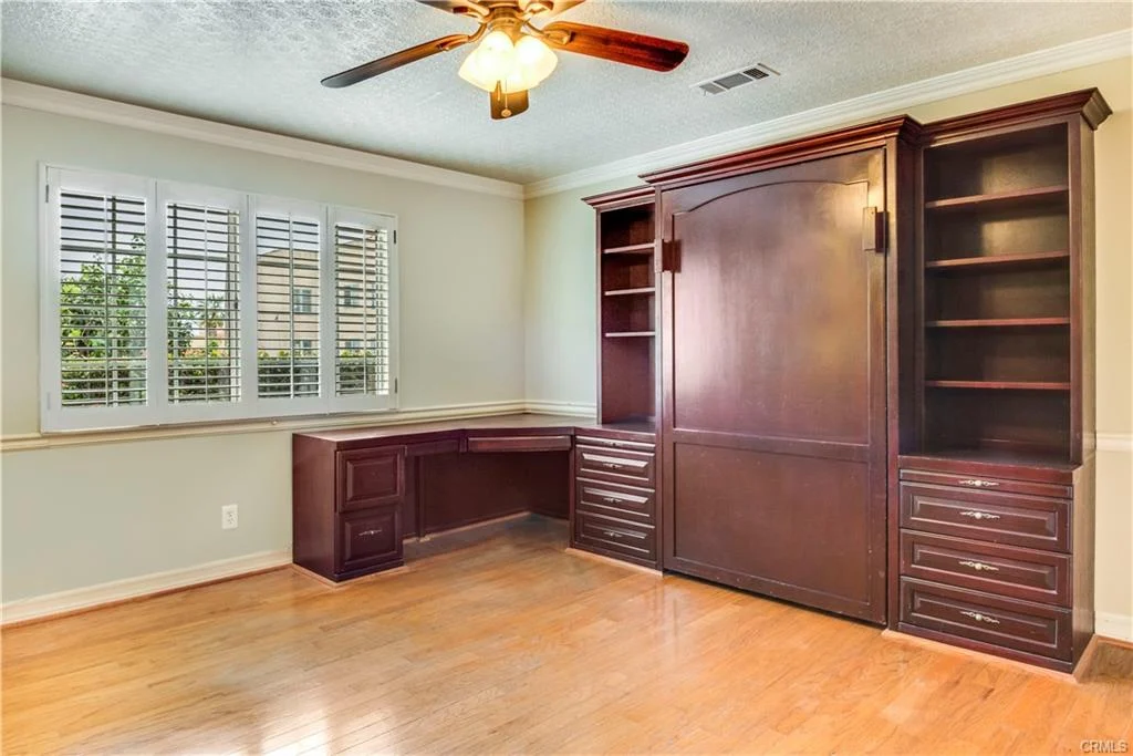 Room with hardwood floor, built-in desk, shelves, ceiling fan, and window with shutters