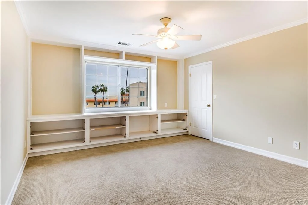 Empty room with beige walls, carpet floor, ceiling fan, window with palm trees, and built-in shelves