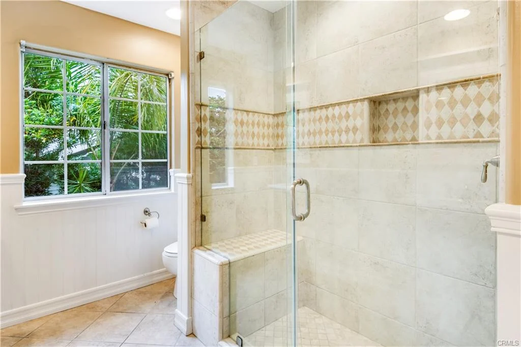 Bathroom with glass shower, beige tiles, window with plants view, and toilet