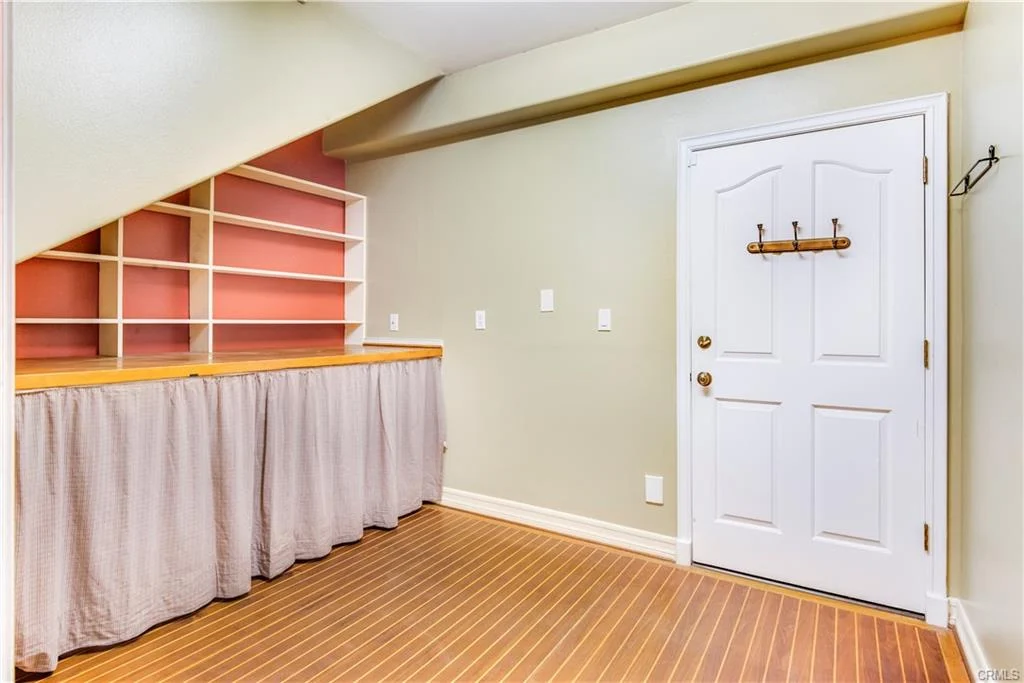 Room with wooden floor, door, wall shelf, curtain below shelf, and hook on wall