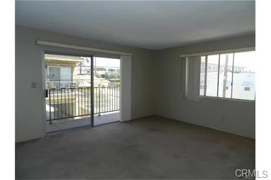 Empty room with beige walls, carpet floor, glass sliding door to balcony, and two windows.