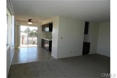 Empty living space with ceiling fan, glass sliding door to balcony, and kitchen area.