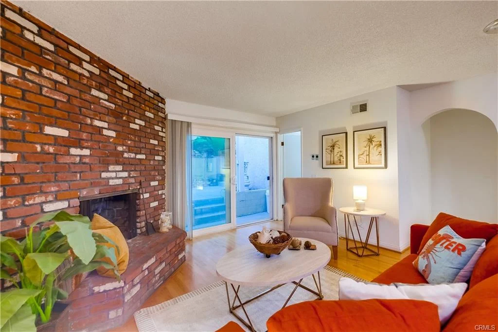 Living room with brick fireplace, red sofa, large windows, plants, and wall art.