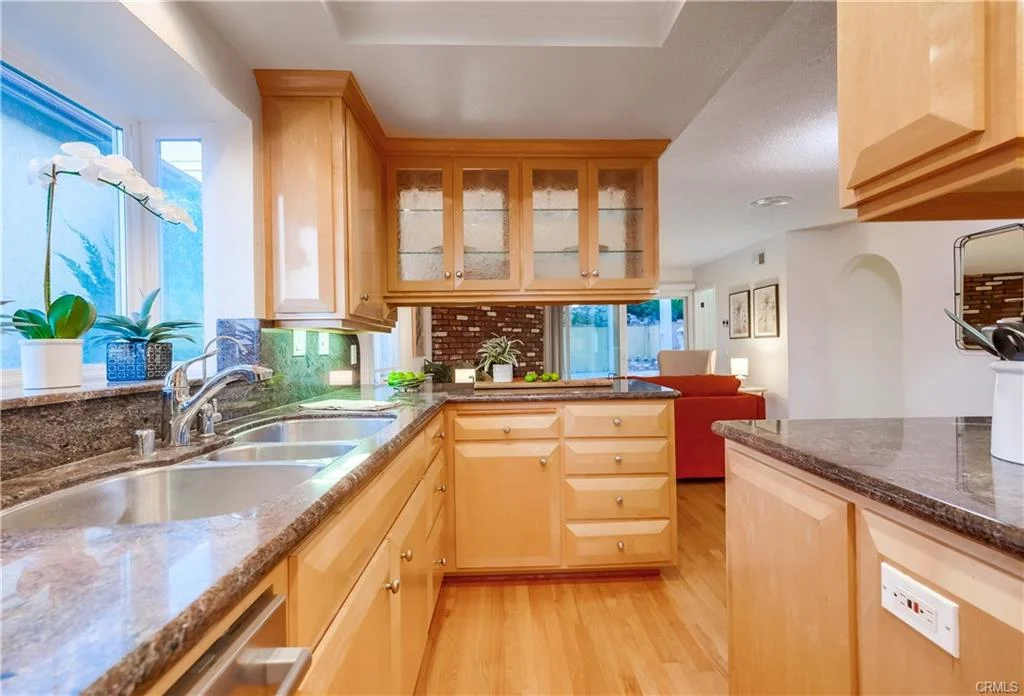 Kitchen with wooden cabinets, double sink, granite countertops, window, and plants.