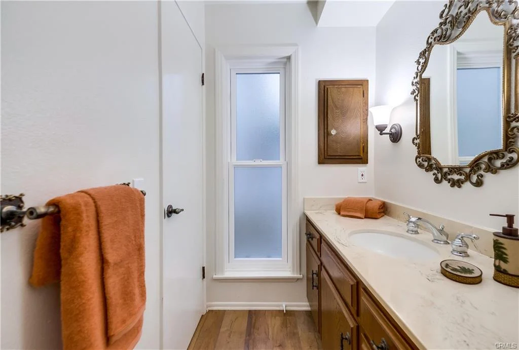 A bathroom featuring a mirror with an ornate frame, a double sink vanity with wooden cabinetry, a window with frosted glass, and an orange towel hanging from the sink.
