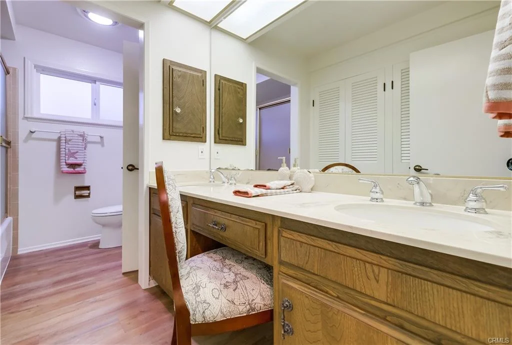 Bathroom with double sink vanity, wooden cabinets, chair, mirror, light walls, wood floor, and separate toilet and shower area.