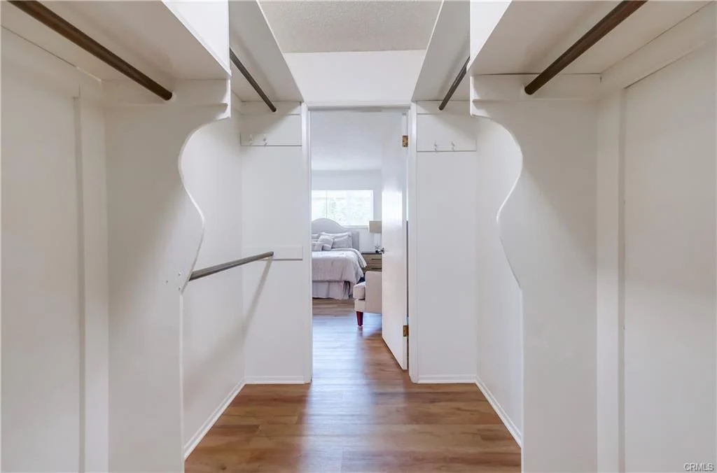 Hallway with two open closets, wooden floor, white walls, leading to room with bed and furnishings.