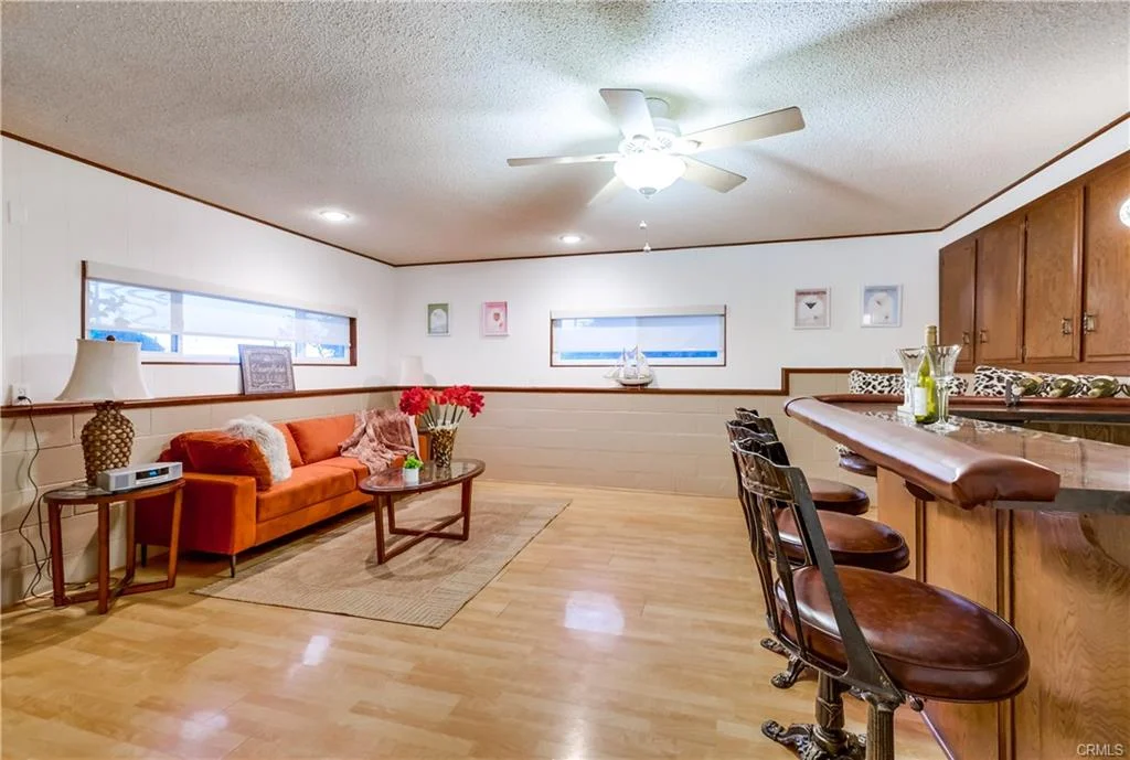 Living room with ceiling fan, orange sofa, glass coffee table, bar with stools, and windows.
