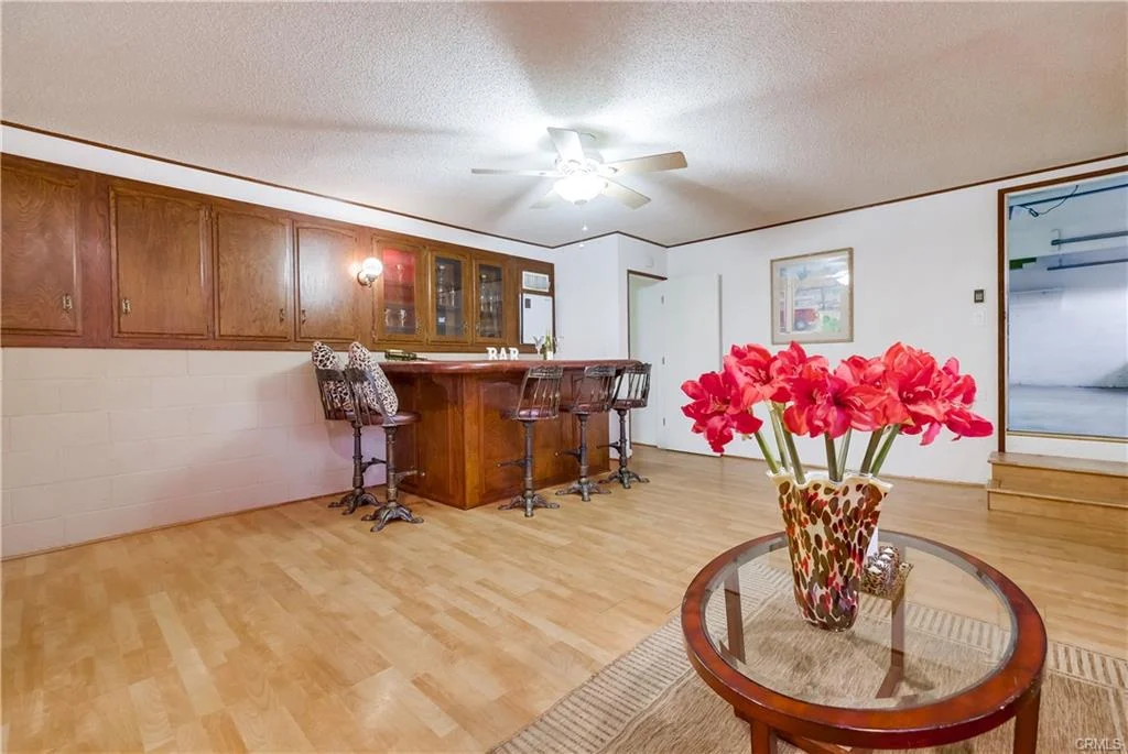 Room with small bar, stools, wooden cabinets, ceiling fan, and vase with pink flowers.