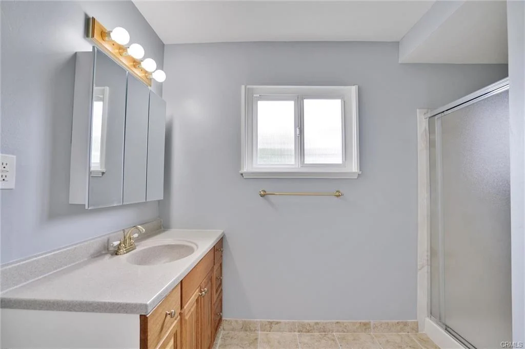 Bathroom with double vanity, wooden cabinets, mirror, window, walk-in shower, light blue walls, and tiled floor.