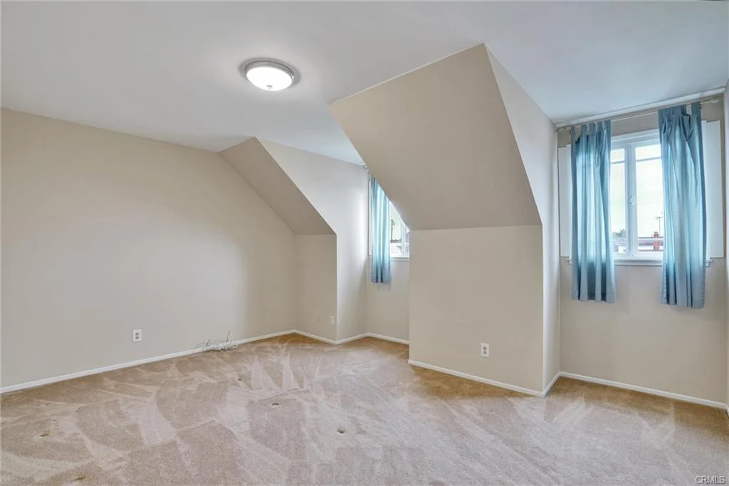 Room with sloped ceilings, light walls, beige carpet, and two windows with blue curtains.