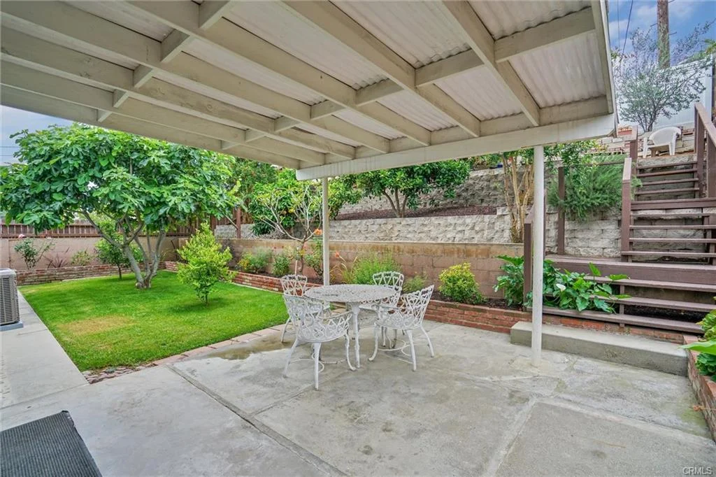 Patio with table and chairs, covered area, green lawn, plants, and stairs.