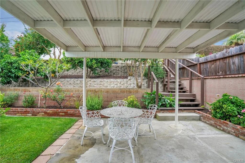 Patio with white table and chairs, covered area, greenery, flowers, and staircase.