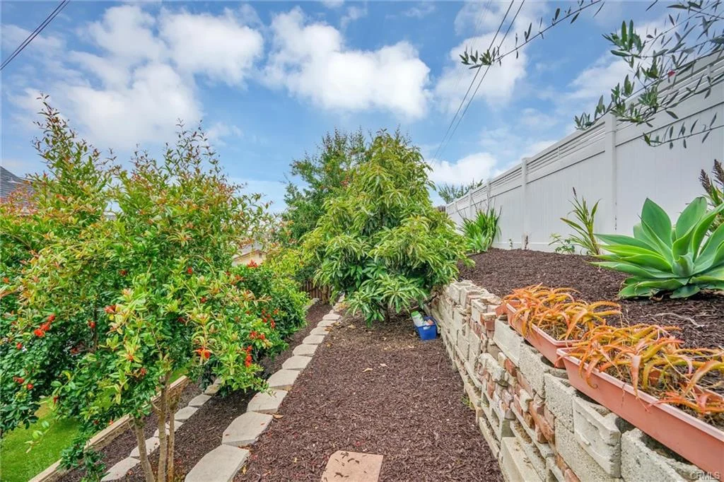 Pathway with plants and trees, leading through garden with white fence.