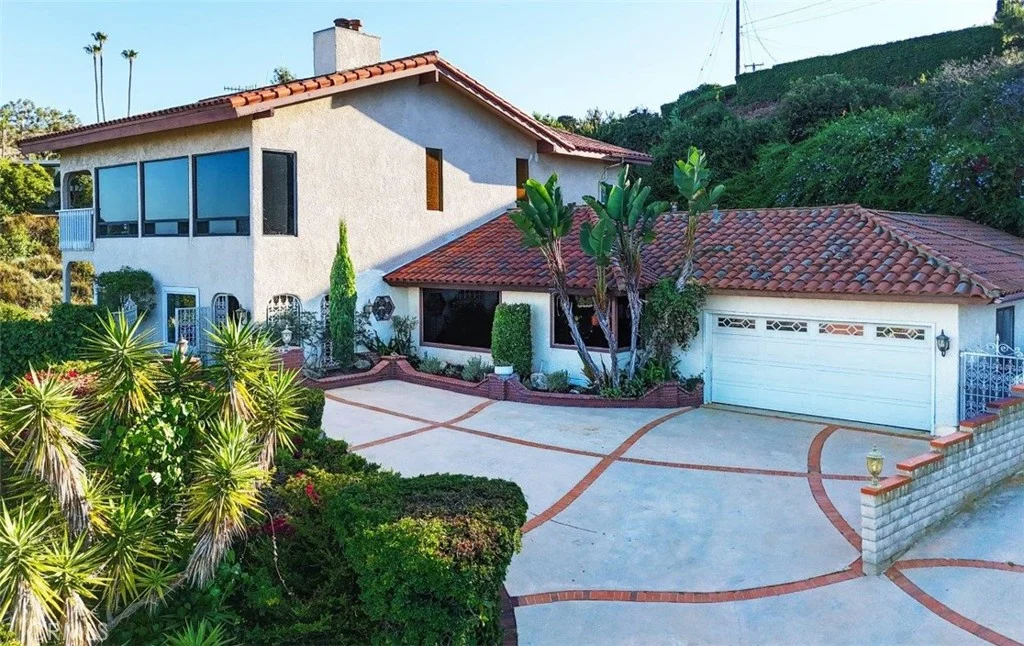 House California real estate with red tile roof, palm trees, green plants, curved driveway, and two car garage