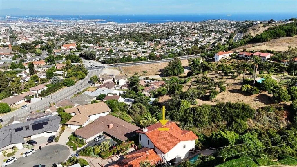 White house California real estate with red tile roof marked by yellow arrow in neighborhood aerial view with green hills