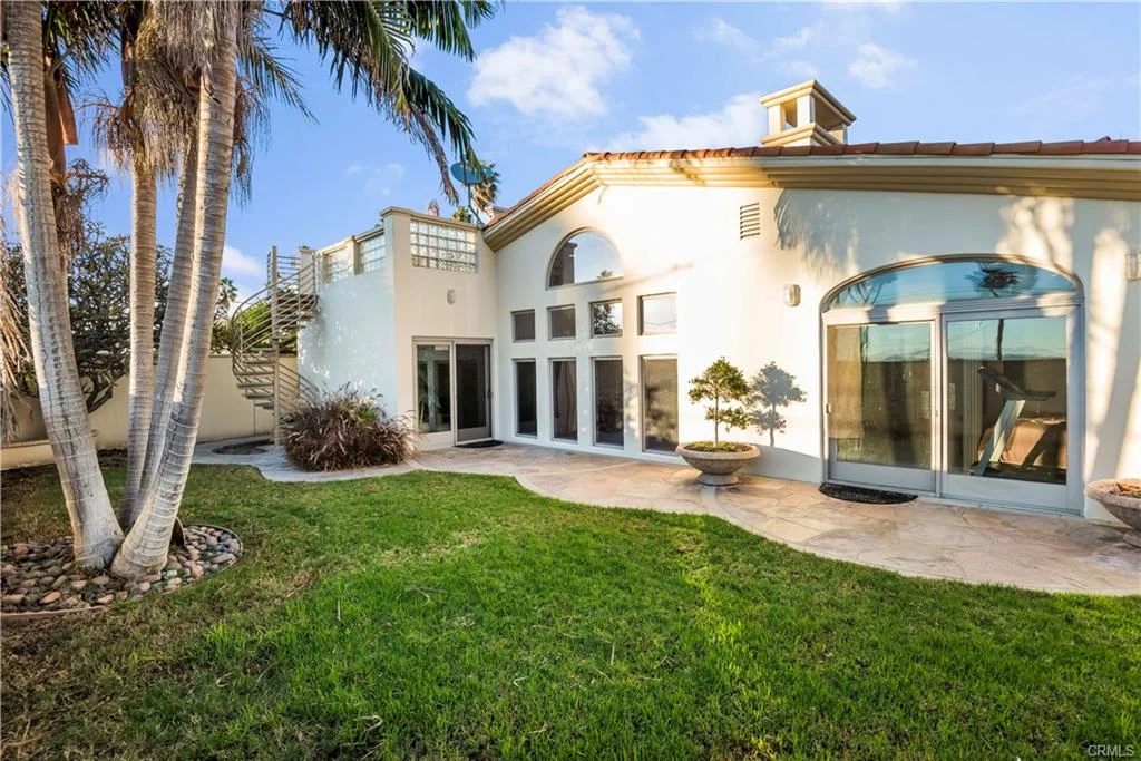 Backyard with green plants, palm trees, patio, large windows, and glass doors.