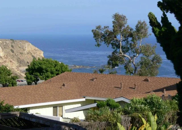 Hillside view with trees, rooftops, ocean waves, and rocky shoreline.