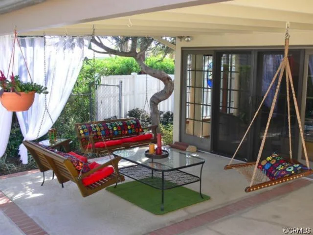 Outdoor seating area with wooden benches, swings, colorful cushions, a glass coffee table, and greenery.