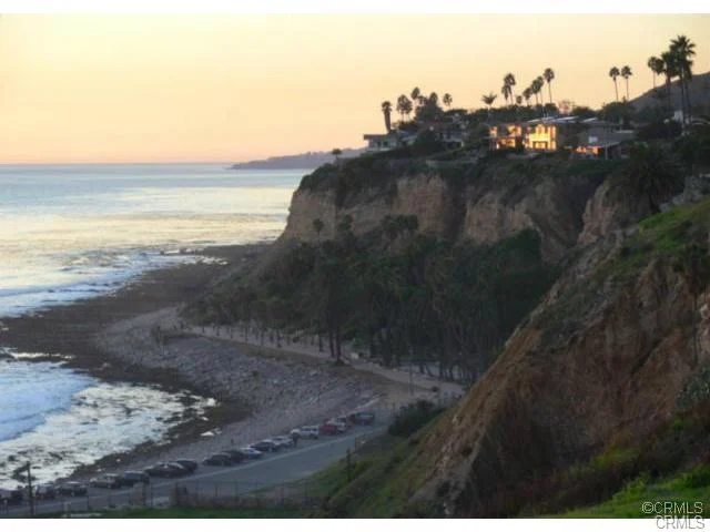 Coastal view at sunset with cliffs, palm trees, a beach, and parked cars.