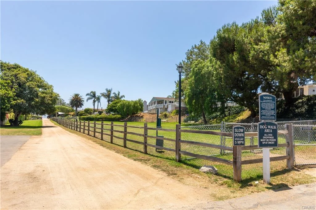 Dirt path leading to park with fence, palm trees, and signs for dog walking area