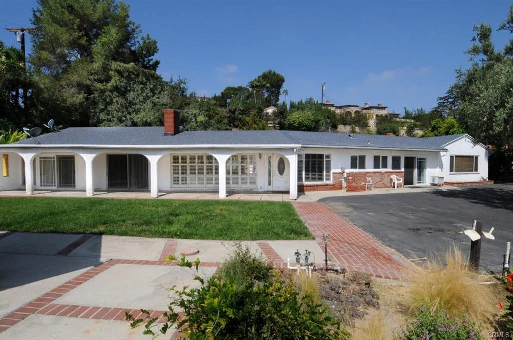 California real estate house with red brick chimney, windows, paved driveway, walkway, and green plants with trees.