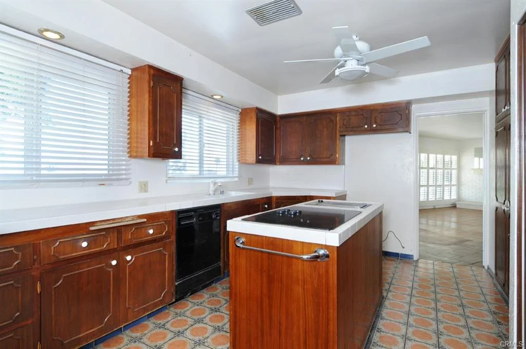 Kitchen with brown wooden cabinets, white counter, black dishwasher, ceiling fan, tiled floor, and windows with blinds.