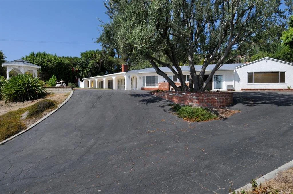 Driveway to California real estate house with white wall, red brick base, and green plants with trees.