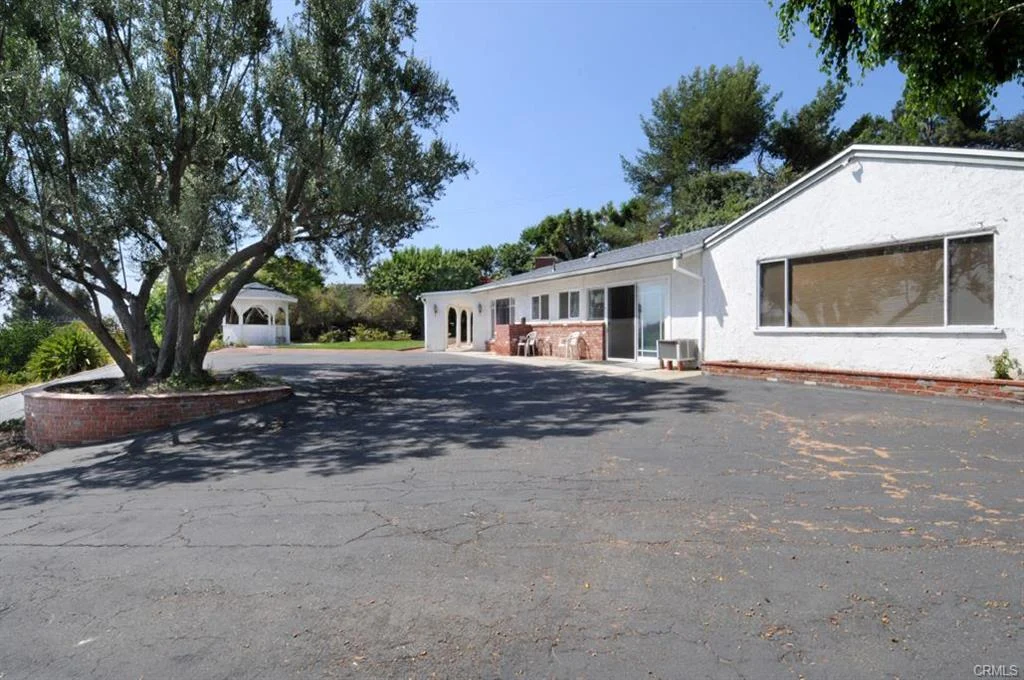 California real estate house with white wall, big driveway, and green plants with trees.