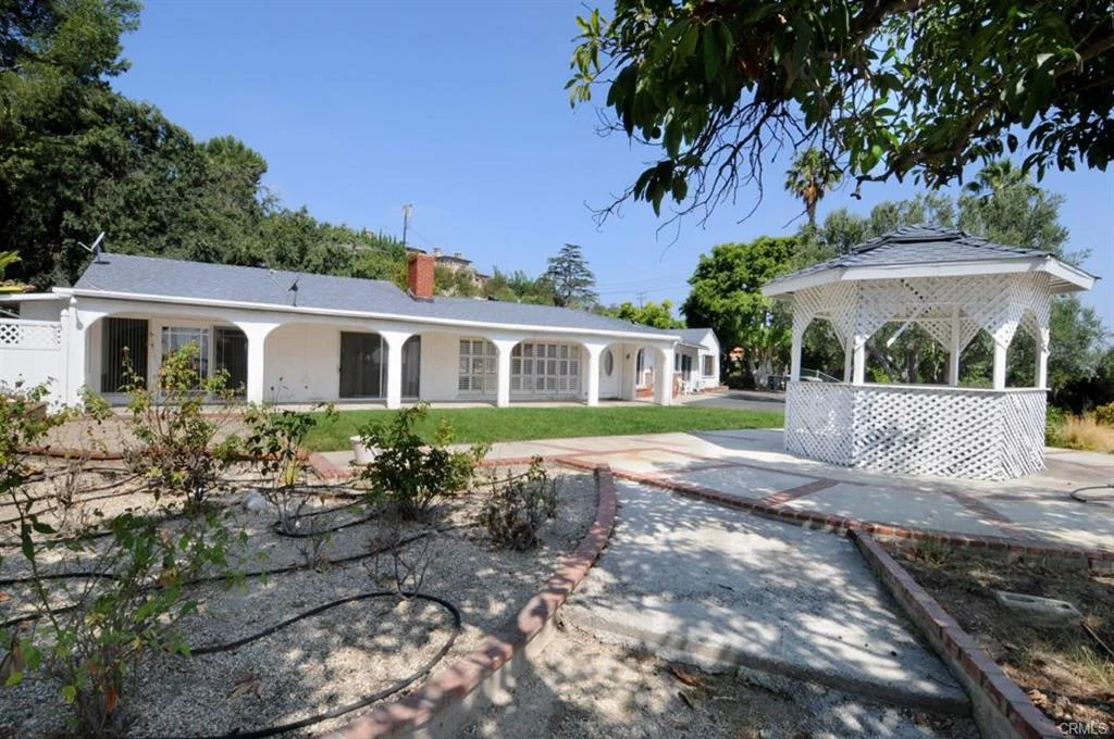 California real estate house with white wall, gazebo, and green plants with trees.