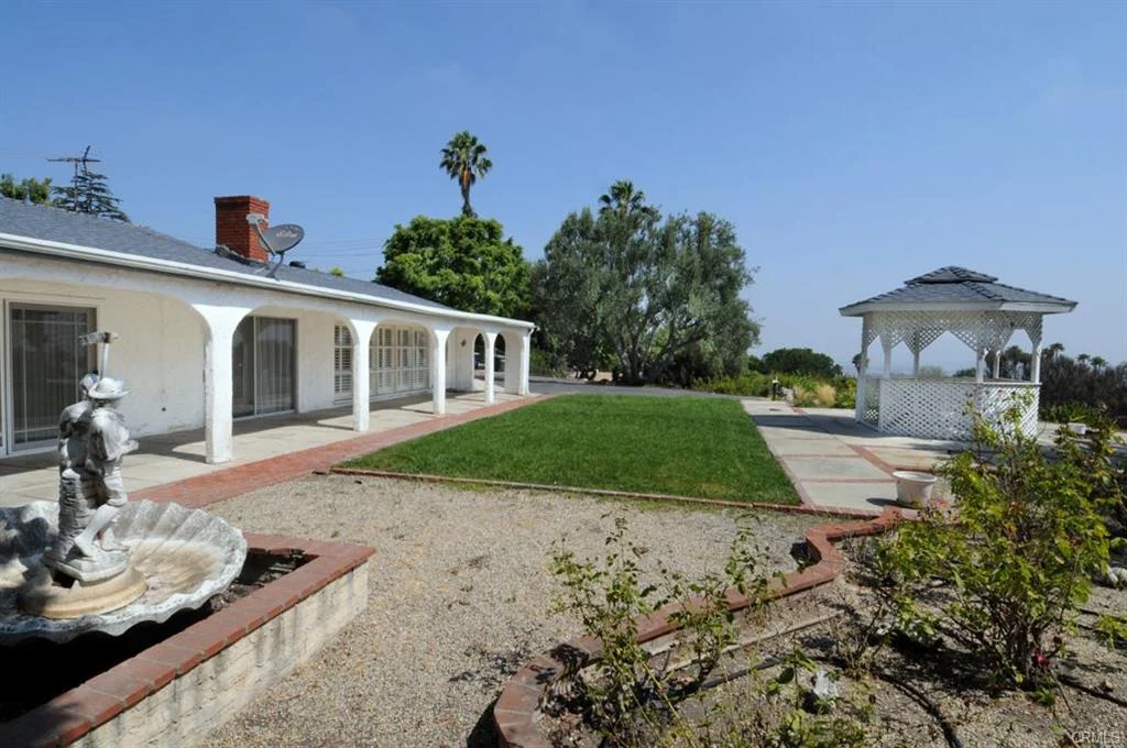 Yard with white gazebo, stone fountain with statue, white building with arched windows, palm trees, and green plants.
