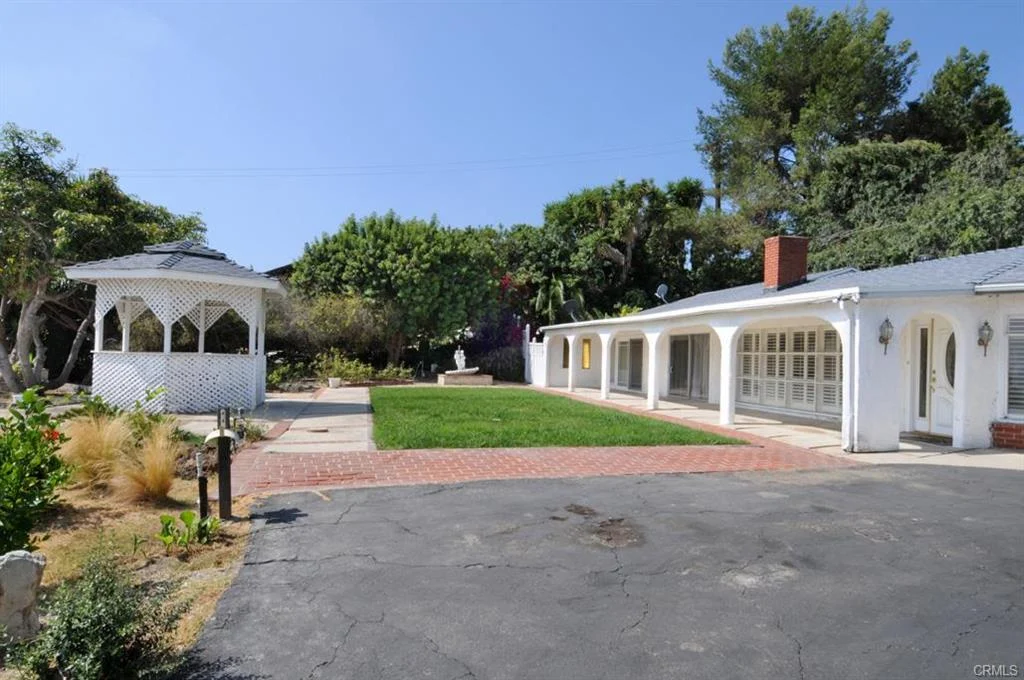 Luxury white California real estate house with green plants lawn, gazebo, black driveway, and trees.