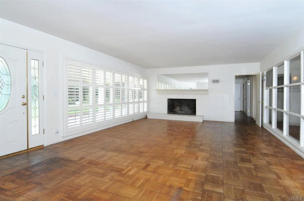 Empty living room with fireplace, large windows with shutters, and wood floor.