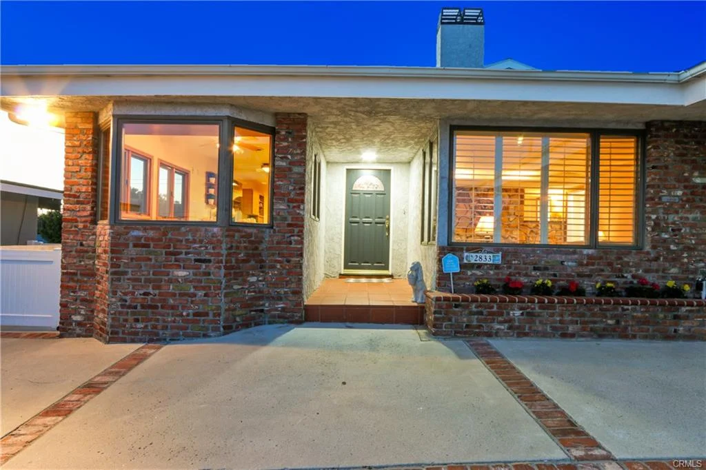 Front view of house with brick walls, porch, black door, windows, and flower beds, California real estate.