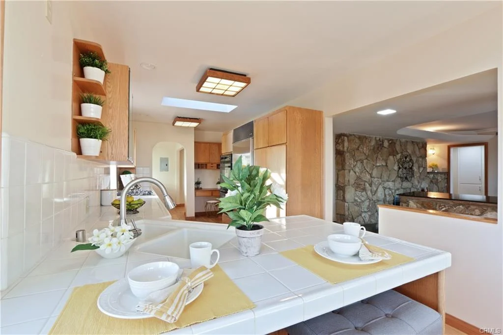 Kitchen with white countertop, wooden cabinets, shelves, and plant.