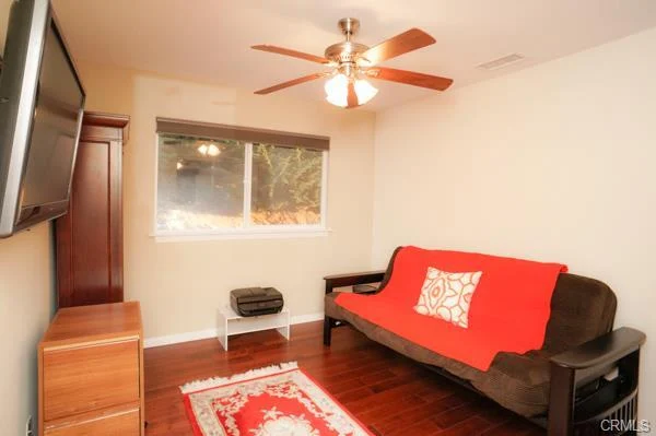 Small room with a couch, a red blanket, a wall-mounted TV, hardwood floors, and a window with blinds.