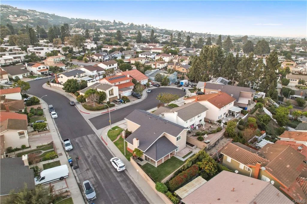 Aerial view of a suburban neighborhood featuring various houses, trees, and a street lined with parked cars.