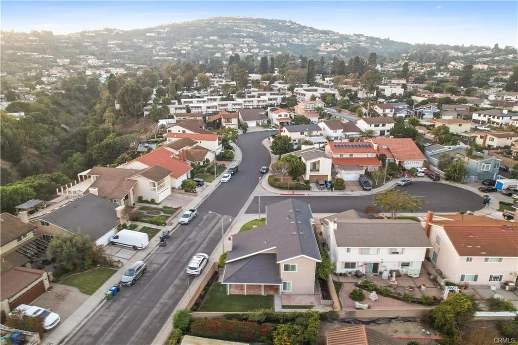 Aerial view of a suburban neighborhood featuring various houses, trees, and a street lined with parked cars.