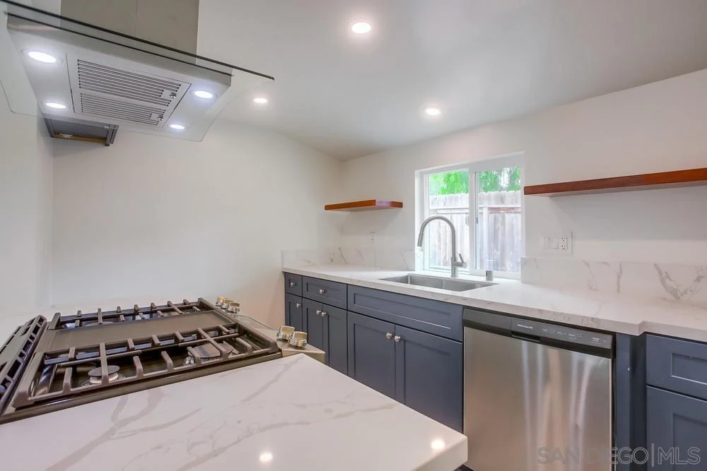Kitchen with gas stove, stainless steel dishwasher, white countertops, wooden shelves, and window above sink.