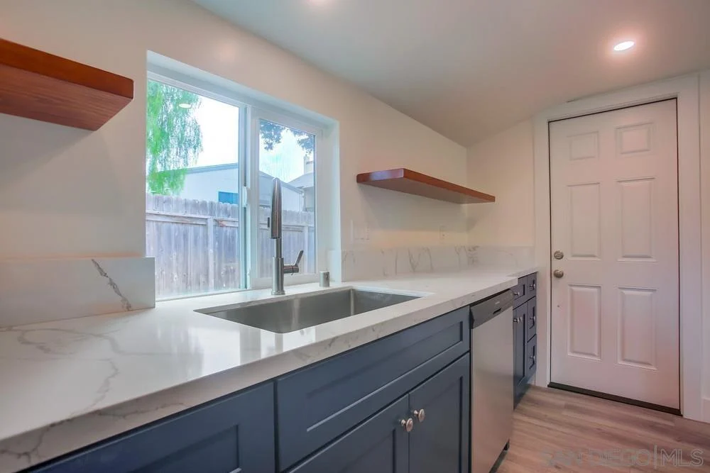 Kitchen with gray cabinets, white countertop, open shelves, sink under window, and door.