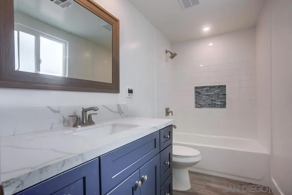 Bathroom with blue vanity, marble countertop, mirror, and tub with tile accent.