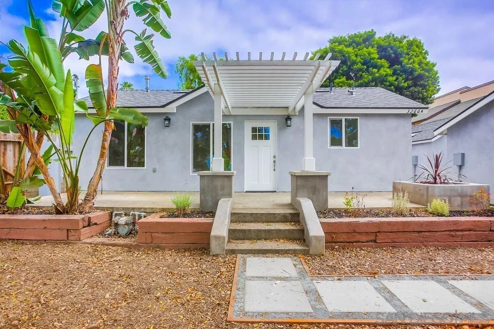 House California real estate with grey exterior, white door, tropical plants, stone pathway, and wooden fence.