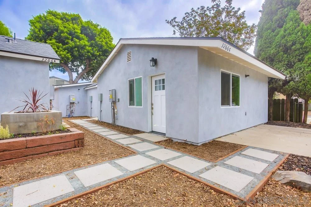 California real estate house with light gray exterior, sloped roof, white trim, green windows, concrete tile path, gravel landscaping, and nearby trees.