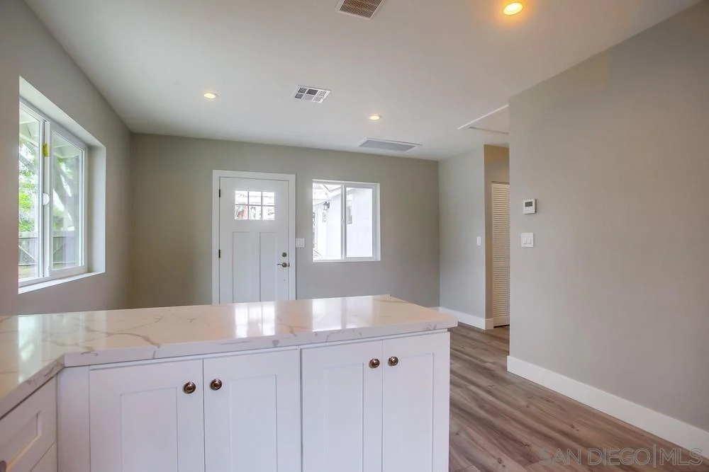 Room with light gray walls, hardwood floor, white kitchen island, windows, and entry door.