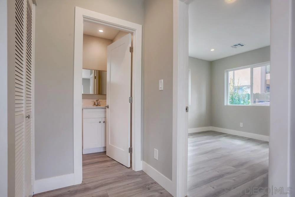 Hallway with light gray walls, wooden floor, bathroom on left, and room with window on right.