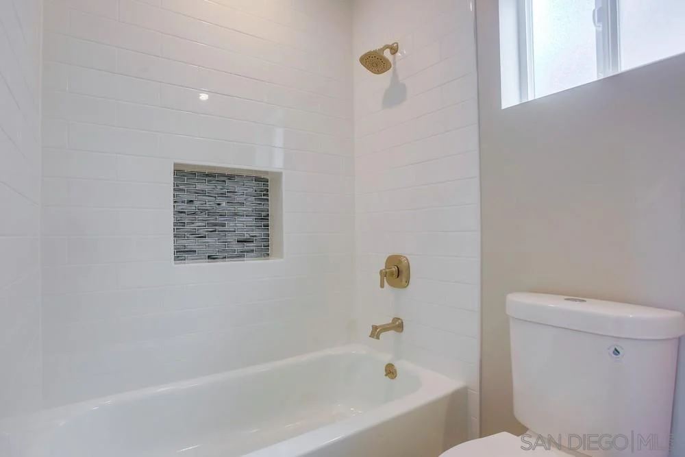 Bathroom with bathtub, showerhead, gold fixtures, white tiles with mosaic, and toilet.
