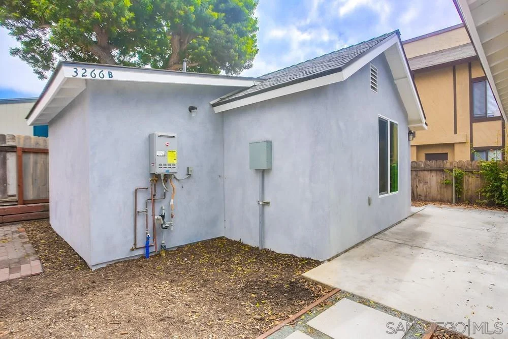 California real estate house with gray exterior, sloped roof, one window, gravel and concrete yard, and utility box.