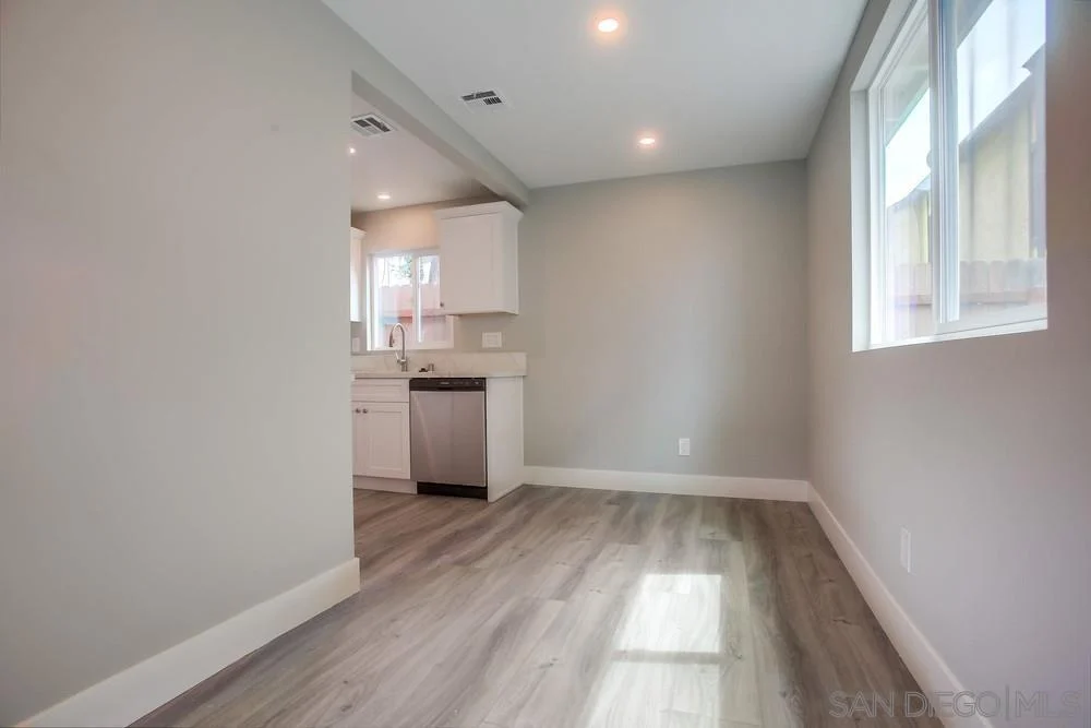 Room with light gray walls, wood-like floor, window, and kitchen with white cabinets.