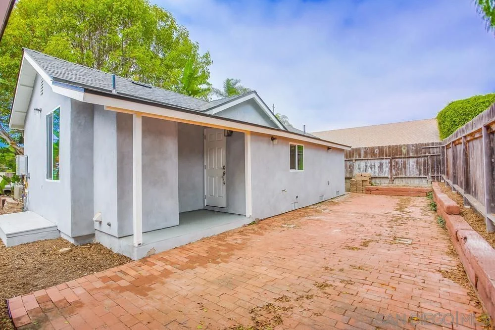 California real estate house with gray exterior, white front door, windows, paved area, and greenery.